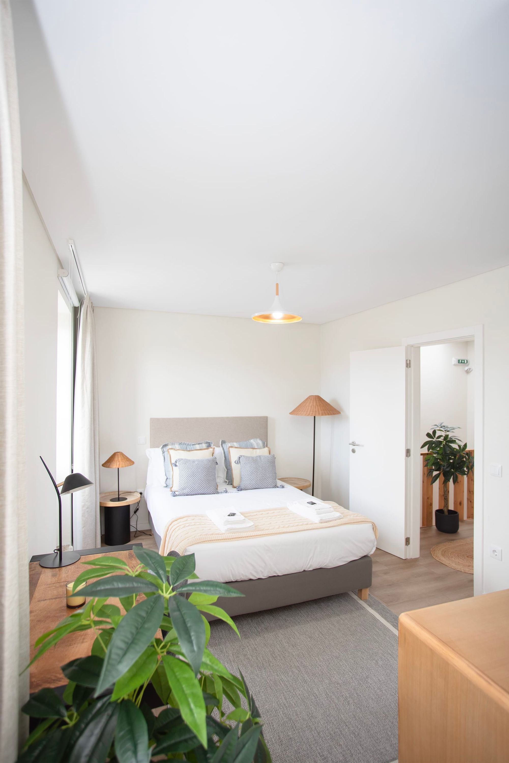 Un dormitorio moderno con cama, lámparas de noche, una mesita de noche de madera, una planta en maceta y vistas a un pasillo.