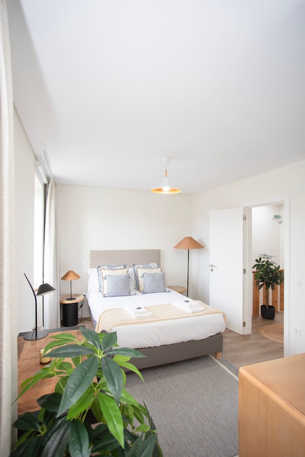 Un dormitorio moderno con cama, lámparas de noche, una mesita de noche de madera, una planta en maceta y vistas a un pasillo.