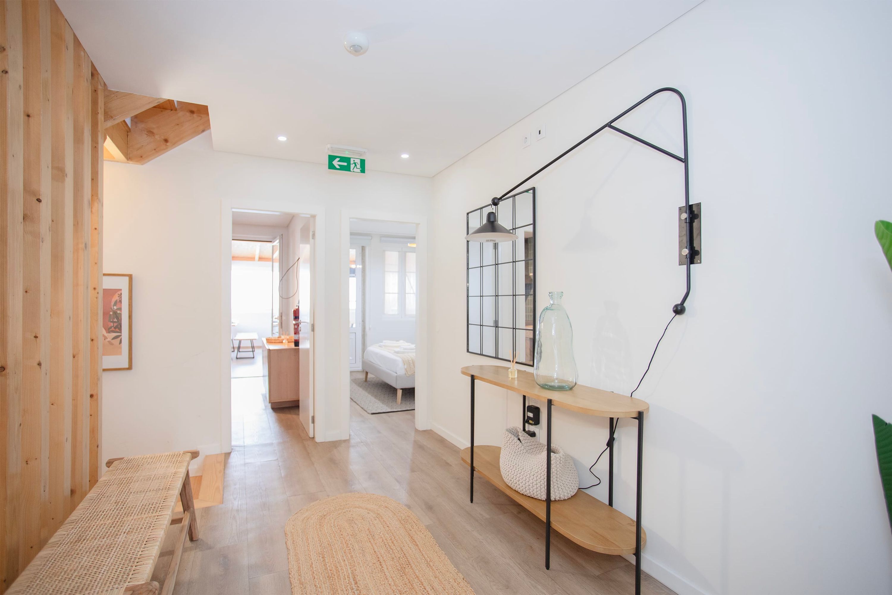 Un couloir d’hôtel avec un banc en bois, une petite table surmontée d’un vase en verre et un miroir accroché au mur.