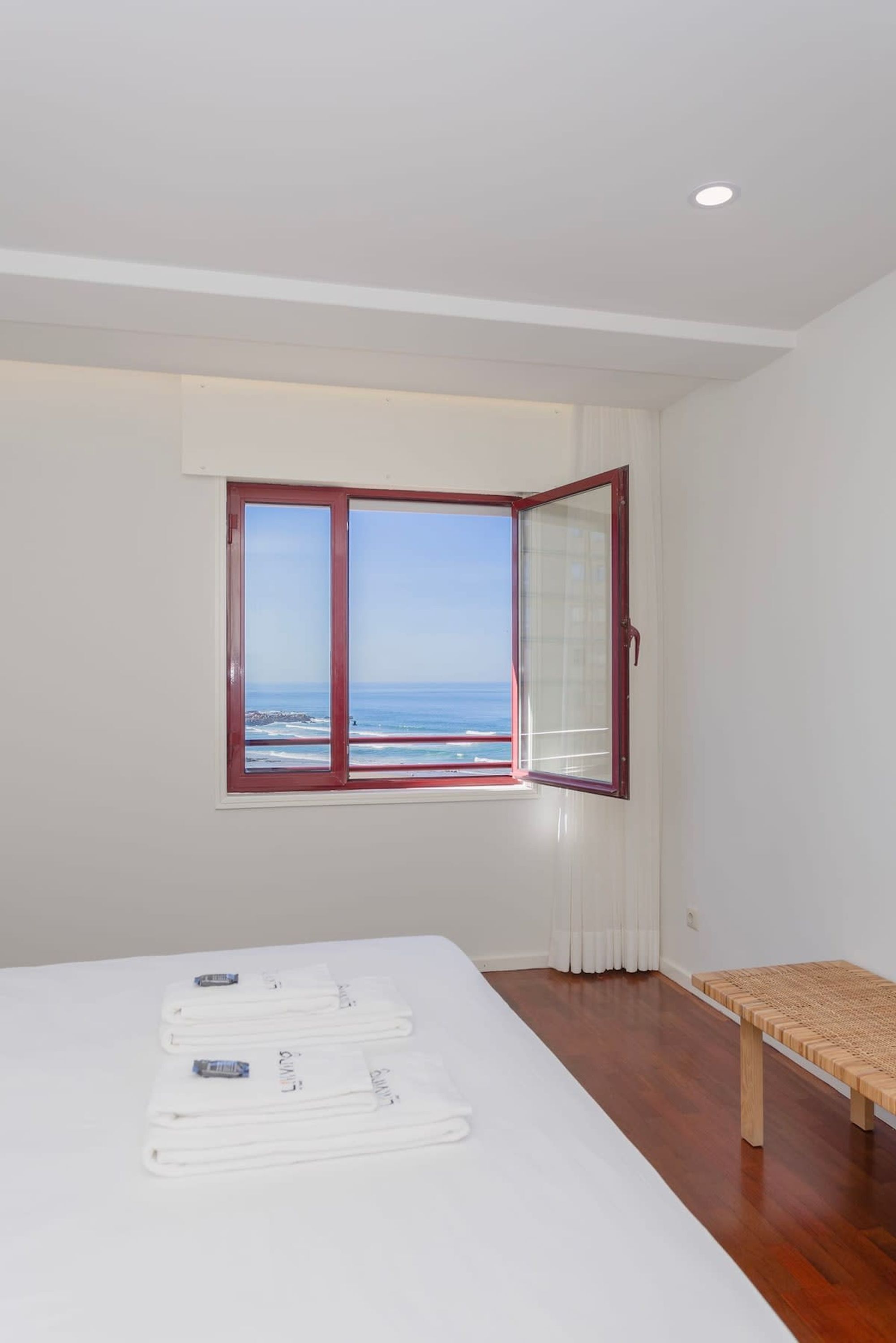 A room with a large window offering a view of the sea, a bed with white linens, and a wooden bench.