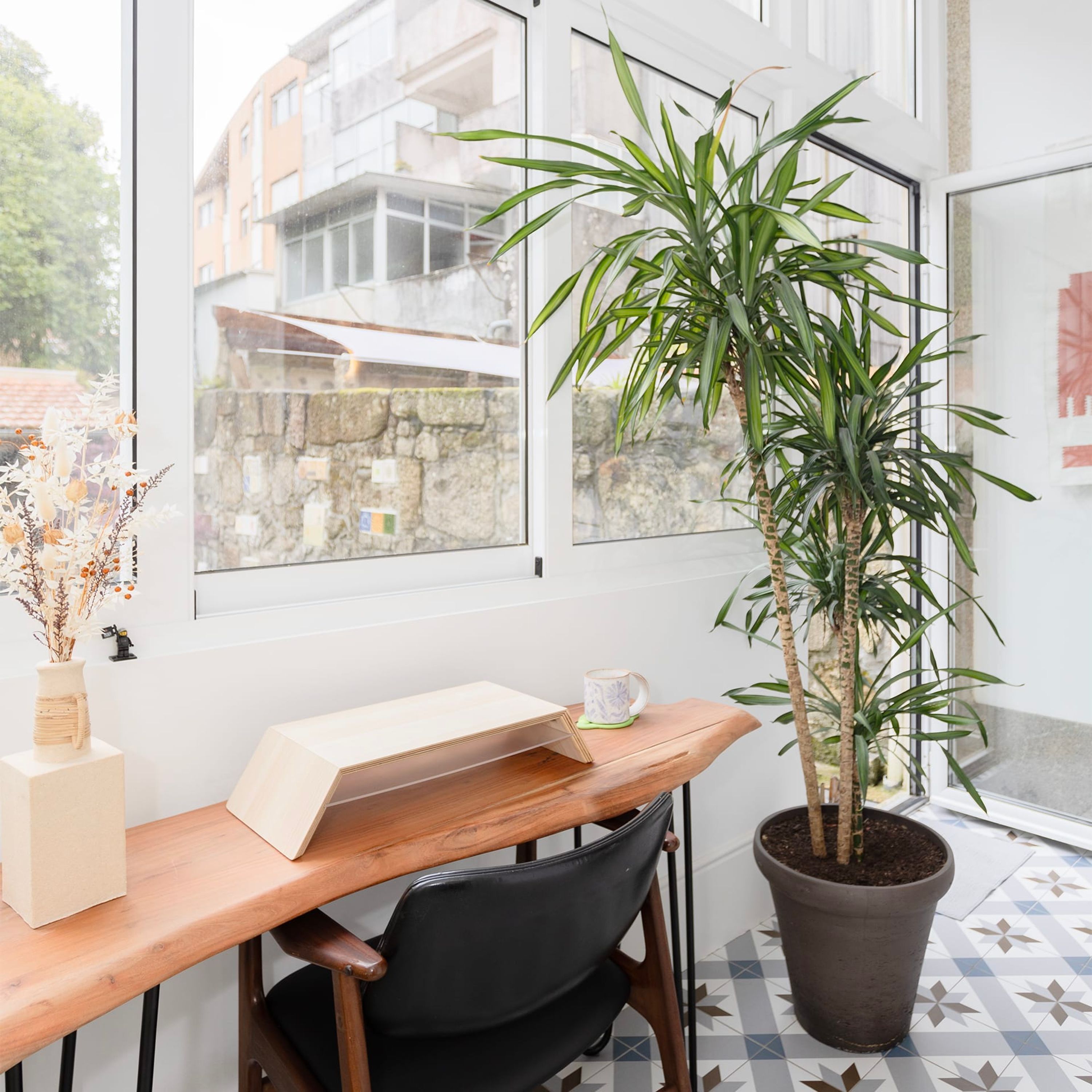 Een kamer met een houten bureau, een zwarte stoel, een plant in pot en een raam met uitzicht op gebouwen.