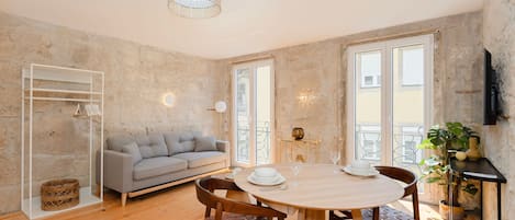 A modern living room with a wooden floor, a grey sofa, a dining table with chairs, and a balcony with a view.