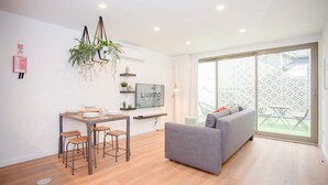 Un salon moderne avec un canapé gris, une table à manger avec des tabourets, un téléviseur fixé au mur et une grande porte-fenêtre coulissante donnant sur un espace extérieur.