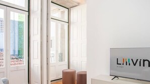 Un salon moderne avec une grande télévision, un canapé et une table basse. On y trouve deux poufs et une porte qui donne sur un balcon.
