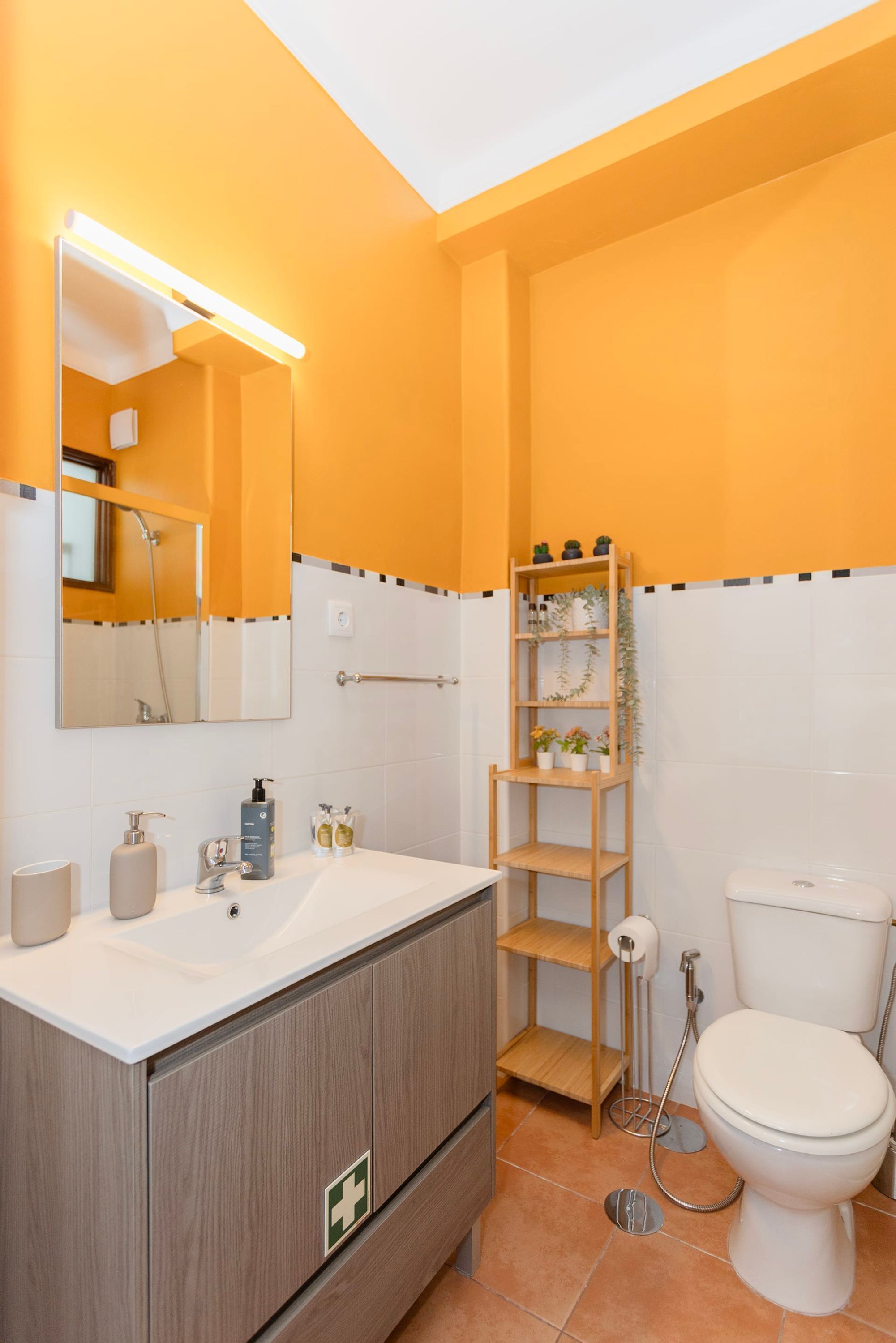 Ein Badezimmer mit weißem Waschbecken, Spiegel und Toilette, Holzregalen mit Toilettenartikeln sowie einer Duschkabine.