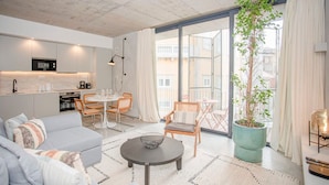 Un appartement moderne comprenant une cuisine, un coin repas et un salon. Il est doté d’une grande fenêtre, d’une plante en pot et d’un coin salon confortable.