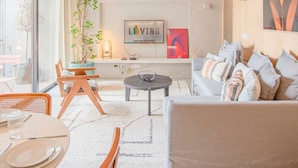 Un salon moderne avec un canapé, une table basse et un coin repas. On y trouve une grande fenêtre, une plante et des œuvres d’art accrochées au mur.