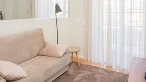 Un salon moderne avec un canapé beige, une petite table ronde, un lampadaire et des rideaux légers.