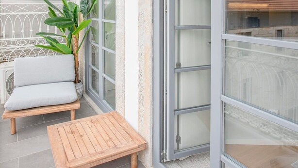 Un balcon avec une table en bois et une chaise rembourrée, un pot de fleurs et une vue sur le paysage urbain à travers de grandes fenêtres.