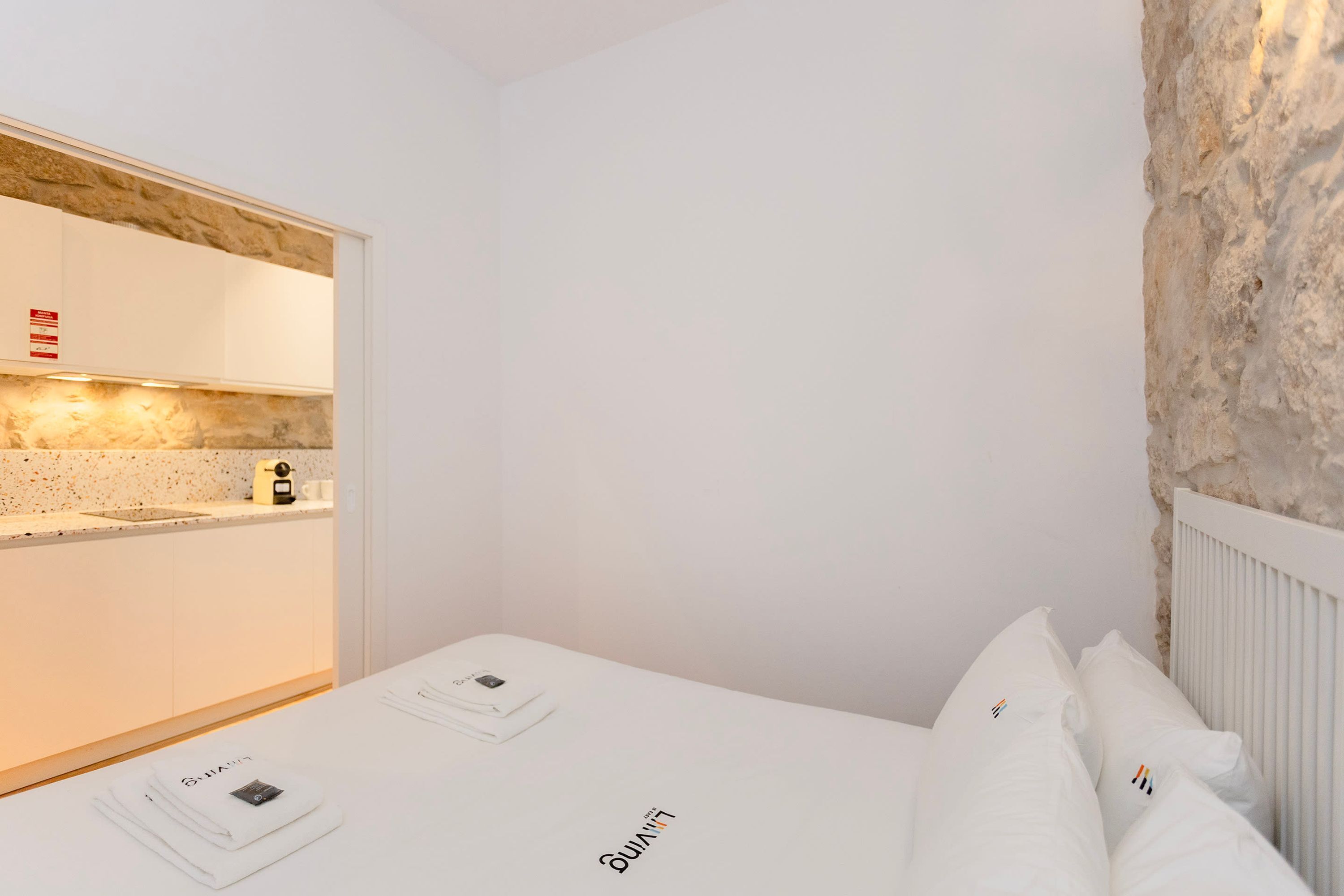 Une chambre d’hôtel avec un coin cuisine, un lit avec du linge de maison blanc et un mur en pierre.