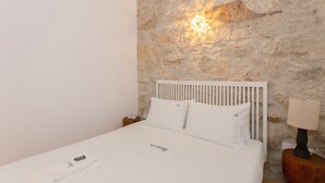 Ein Bett mit weißer Bettwäsche und einem Kopfteil, ein Nachttisch mit Lampe sowie eine Steinwand mit Leuchte.