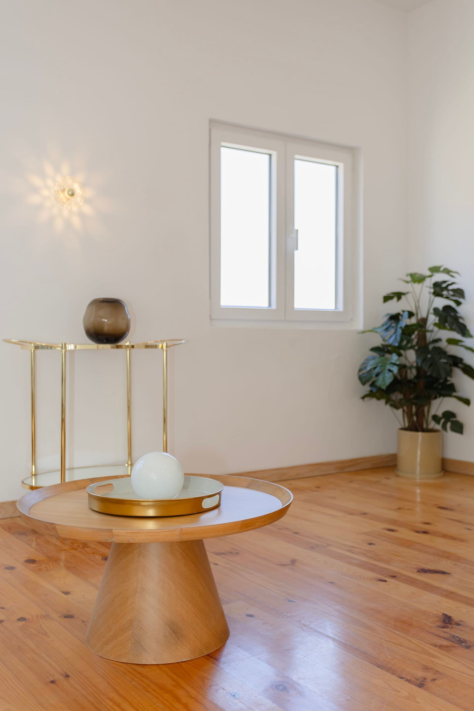 A room with a wooden floor, a round wooden table with a white bowl, a gold vase, and a potted plant.