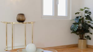 Ein Zimmer mit Holzboden, einem runden Holztisch mit einer weißen Schüssel, einer goldenen Vase und einer Topfpflanze.