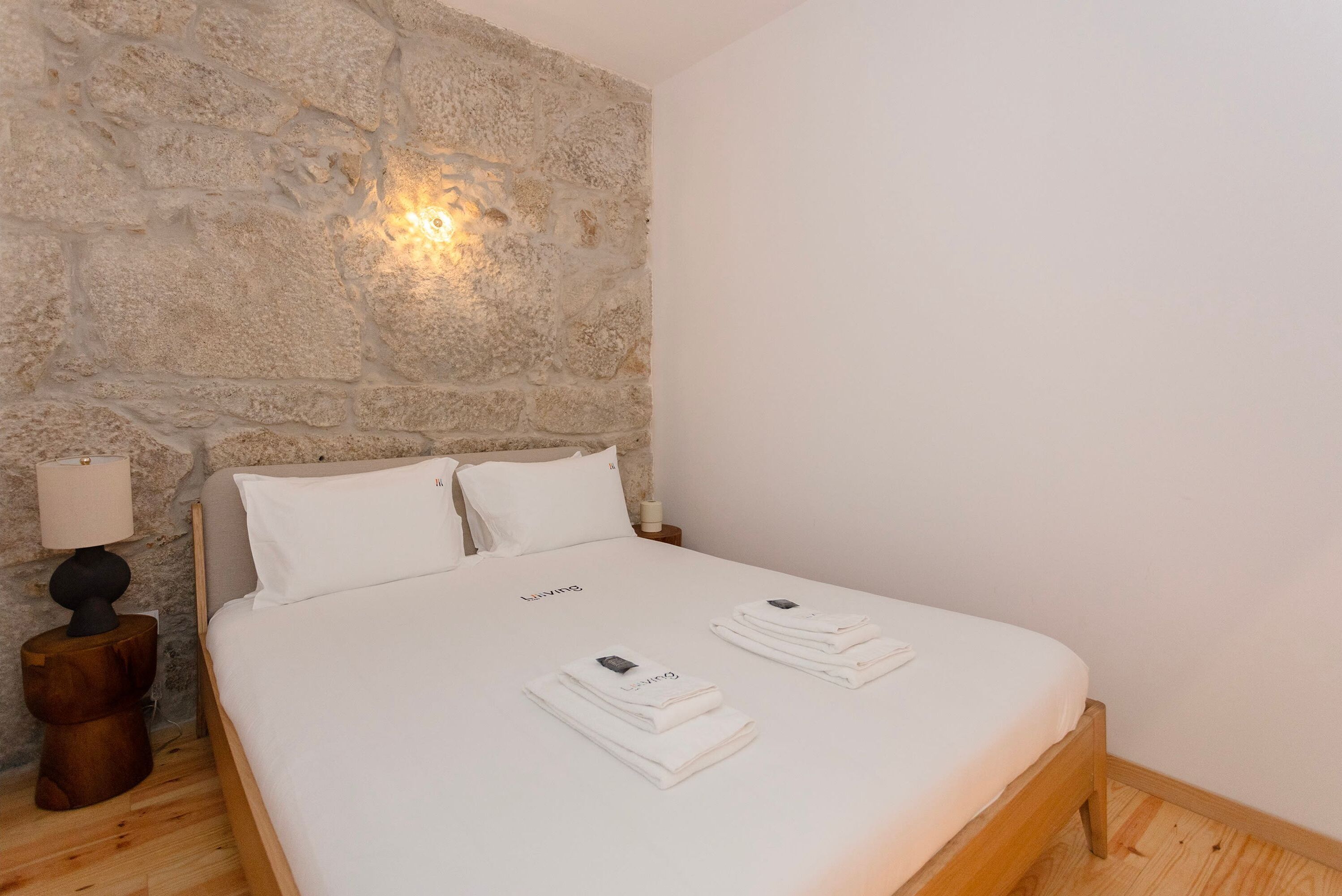 Een netjes opgemaakt bed met witte lakens en handdoeken, een stenen muur en een klein nachtkastje met een lamp.