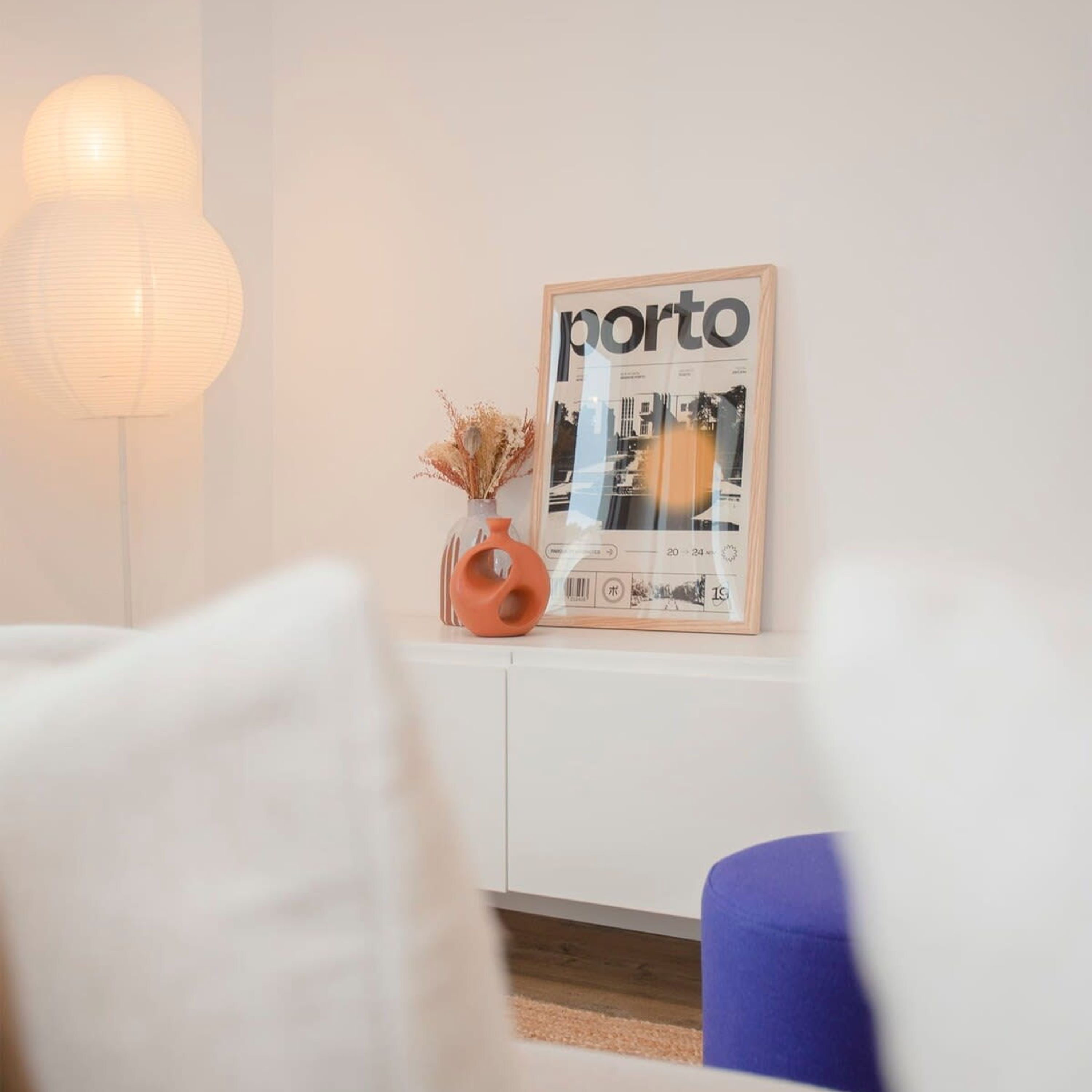 Een kamer met een witte bank, een ingelijst kunstwerk aan de muur, een vaas met gedroogde bloemen en een vloerlamp.