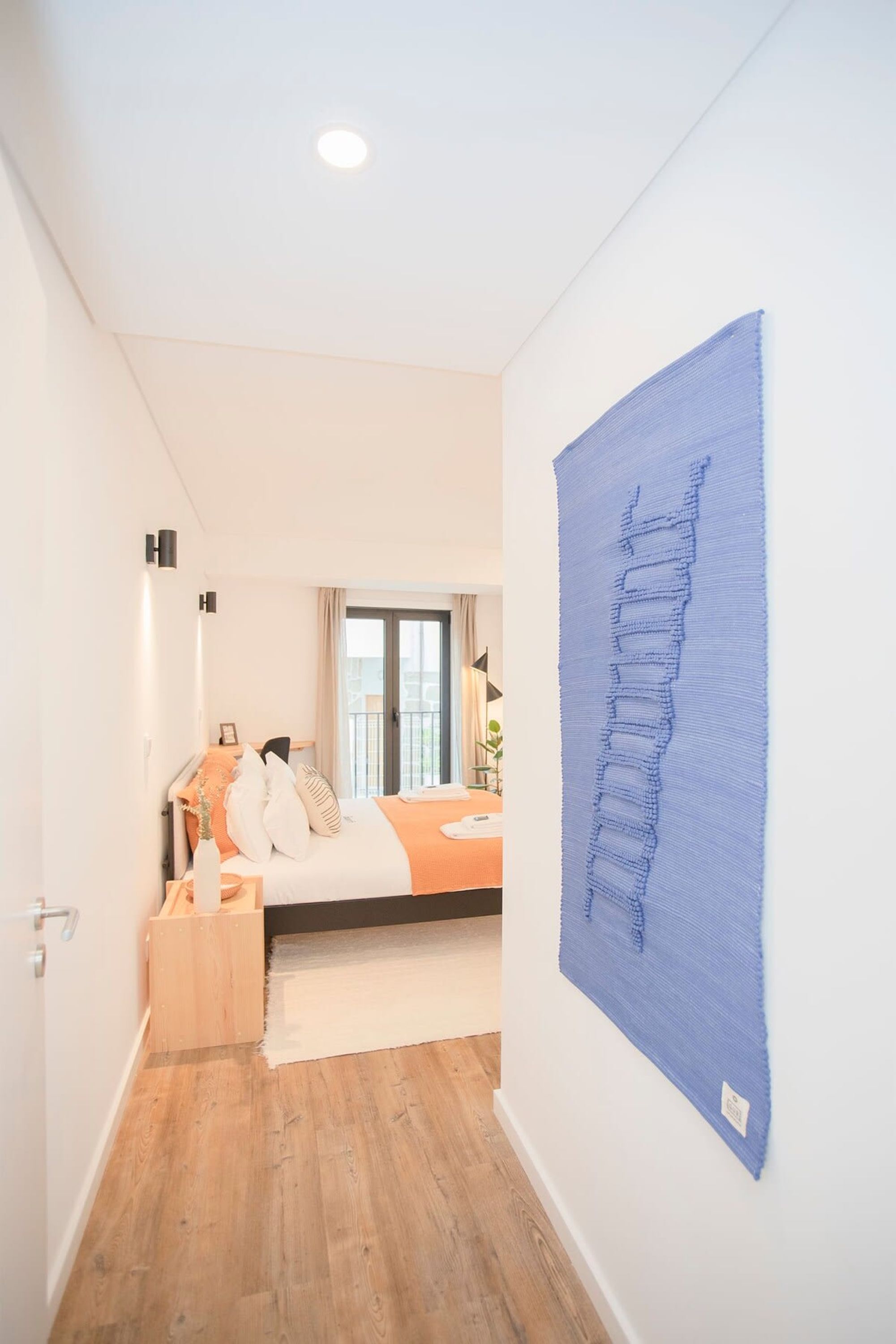 A narrow hallway leading to a bedroom with a bed, a wooden nightstand, and a large blue artwork on the wall.