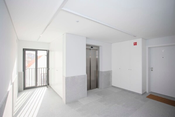 A modern hallway with a glass door leading to a balcony, a central elevator, and a door with a red sign. - Liiiving in Gaia - Cooper Residence Apartment (Vila Nova de Gaia)