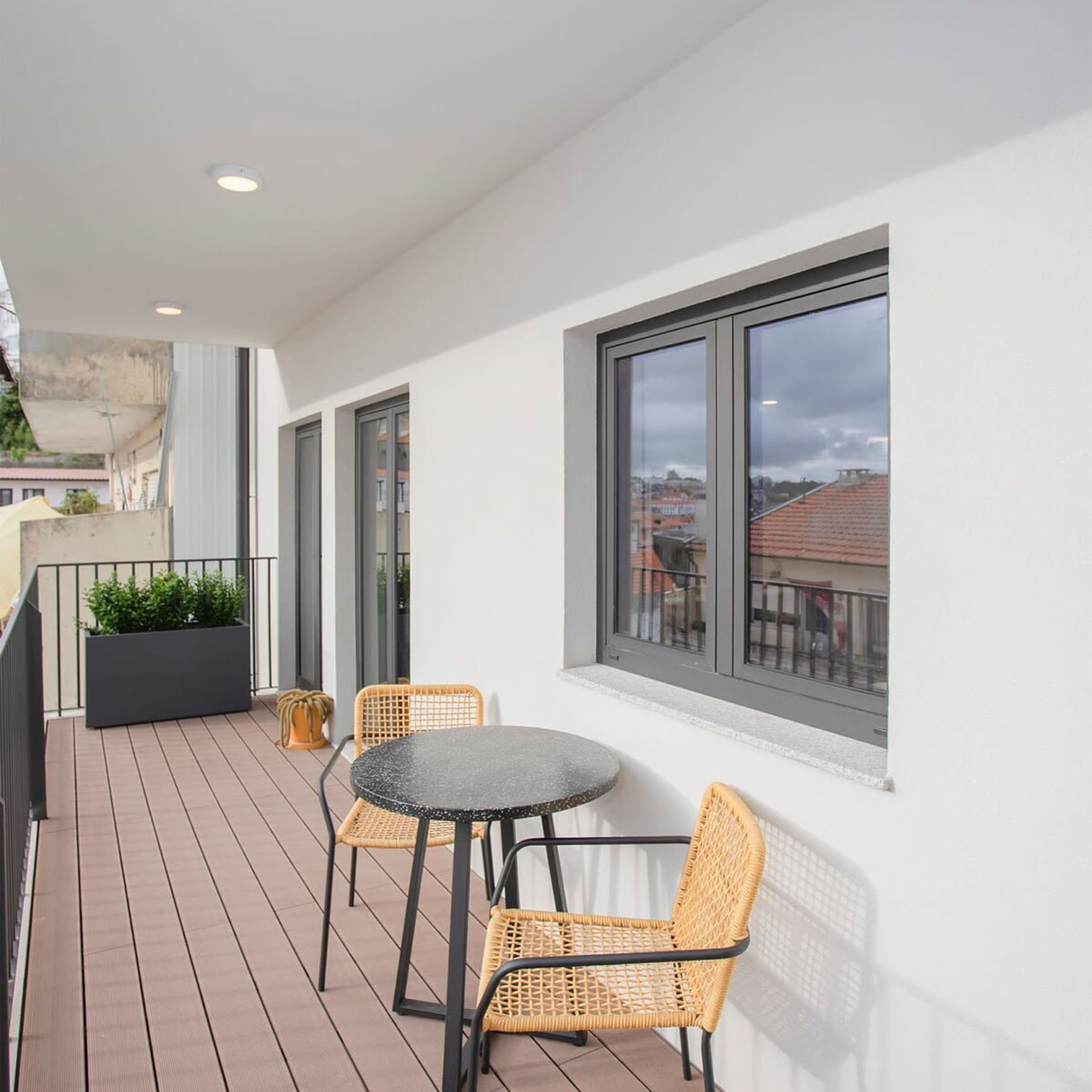 A balcony with a round table and two wicker chairs, a black planter, and a view of the cityscape through large windows.