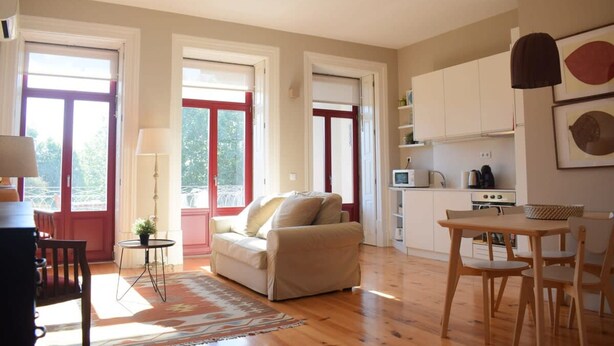 Un salon chaleureux avec un canapé, une petite table et un coin repas avec des chaises. De grandes fenêtres aux cadres rouges, un coin cuisine avec des placards et un ventilateur de plafond complètent l’ensemble.
