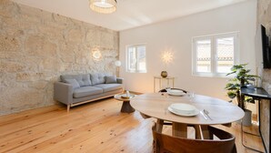 A modern living room with a stone wall, a grey sofa, a wooden dining table with chairs, and a ceiling light fixture. - Liiiving In Gaia - The Wine Side V (Vila Nova de Gaia)