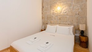 A neatly made bed with white linens and towels, a stone wall with a wall-mounted light, and a wooden nightstand with a lamp. - Liiiving In Gaia - The Wine Side V (Vila Nova de Gaia)