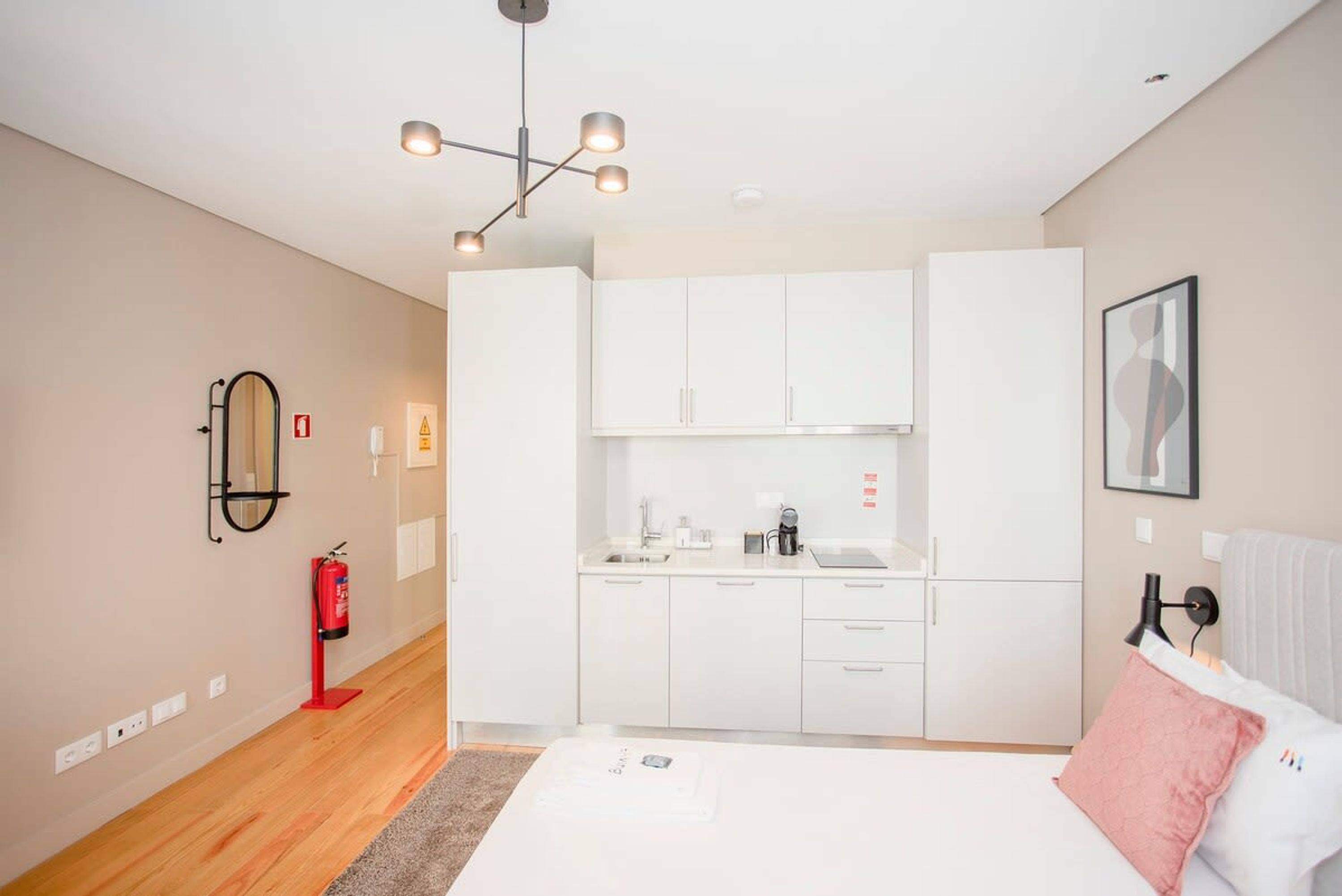 A modern hotel room with a kitchenette, a bed with a pink pillow, a ceiling light fixture, and a fire extinguisher on the wall.
