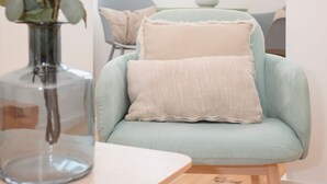 Ein hellblauer Sessel mit einer strukturierten, beige farbenen Sitzkissen, eine transparente Glasvase mit Grünpflanzen und ein Holztisch in einem hellen Raum mit einem Fenster.