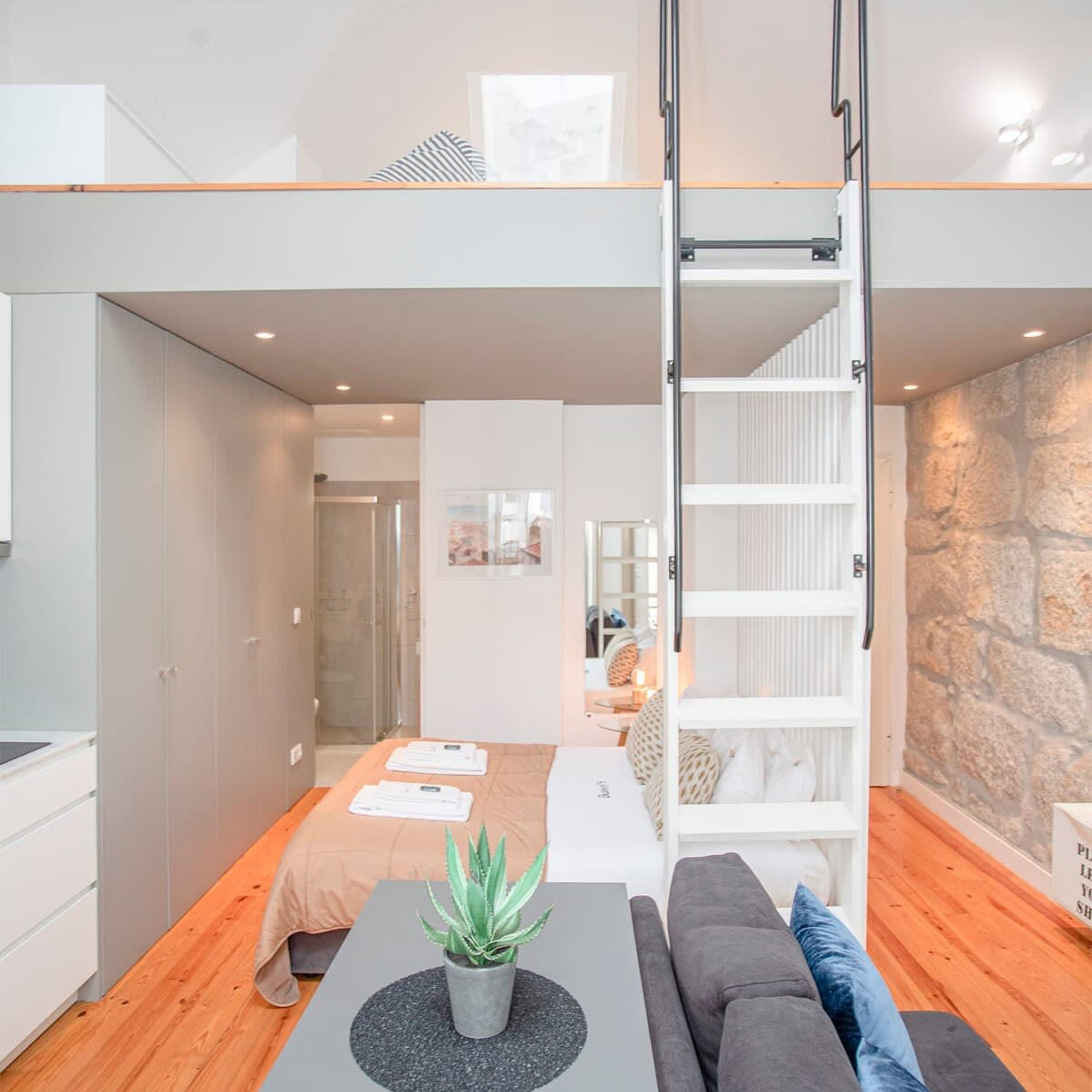 A modern loft apartment with a loft bed, a dining area with a plant, and a stone accent wall.