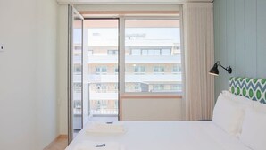 Habitación de hotel con una cama grande, una ventana con vistas a edificios, una mesita de noche y una lámpara.