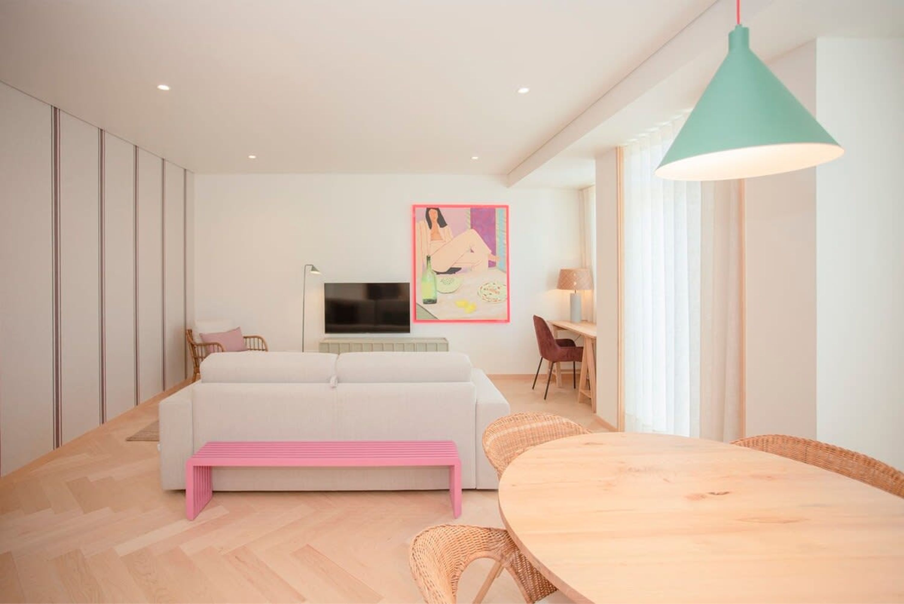 A modern living room with a white sofa, a pink bench, a wooden dining table, wicker chairs, a teal pendant light, a flat-screen TV, and a framed artwork on the wall.