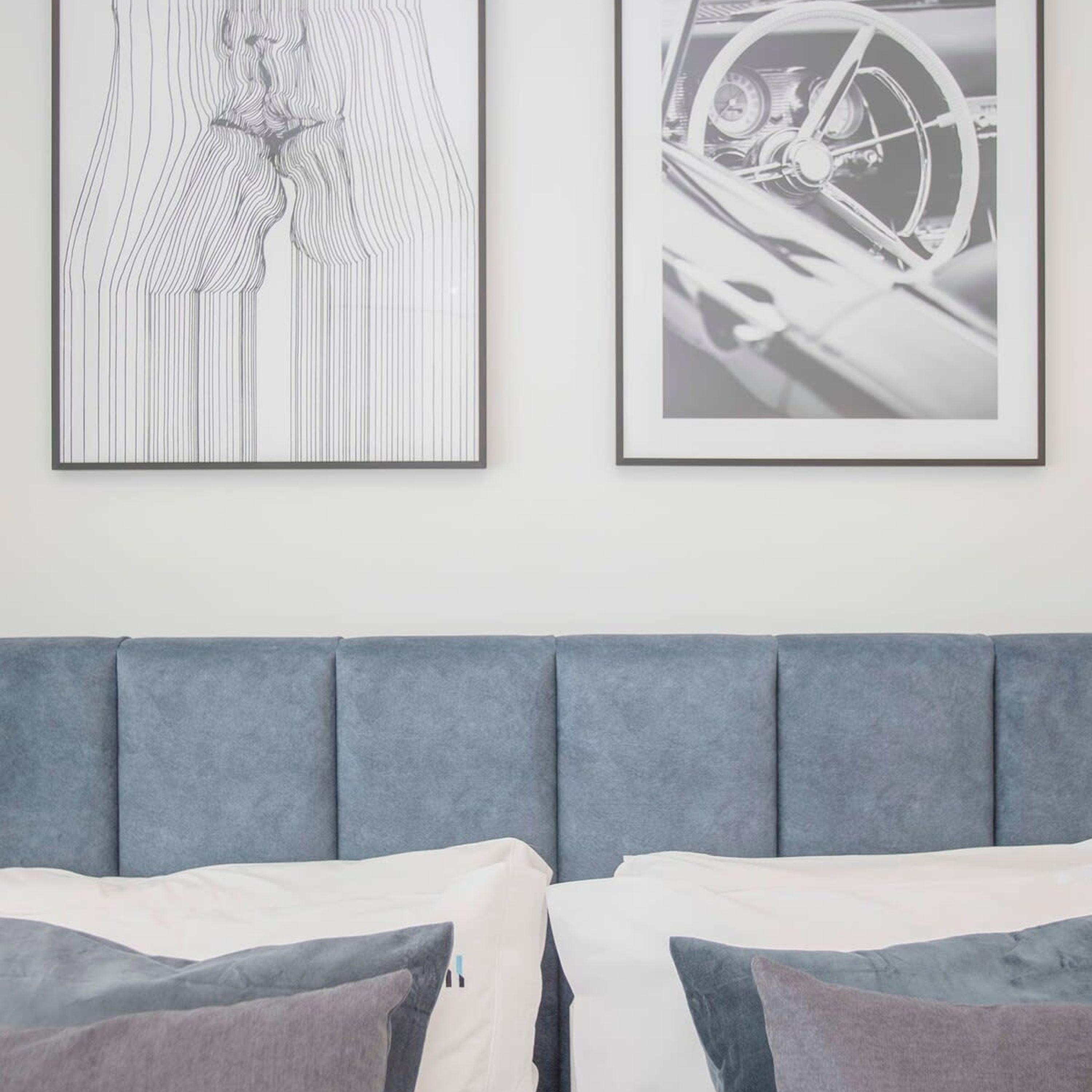 A bedroom with a grey upholstered headboard, a bed with white and grey bedding, and two framed artworks on the wall.