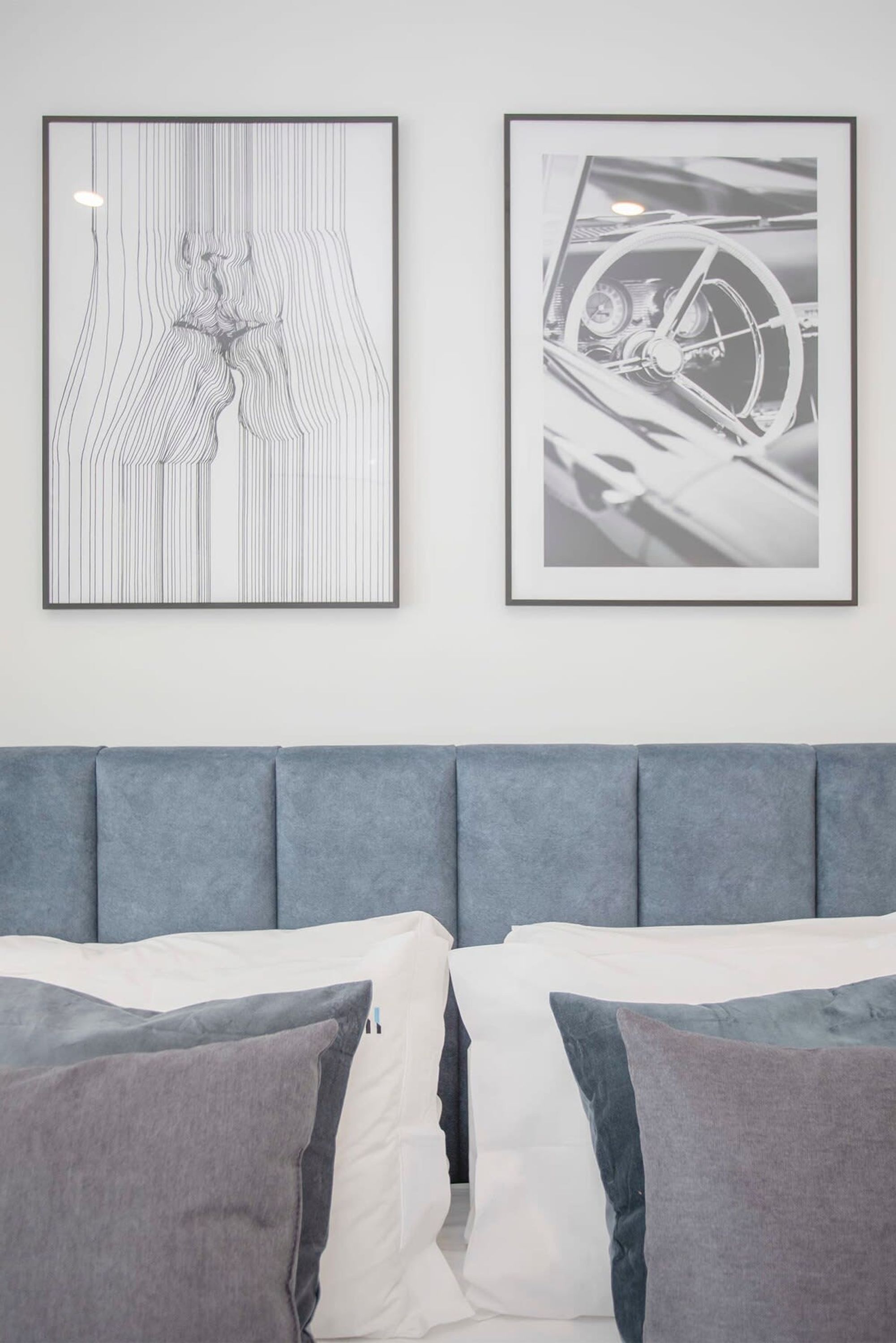A bedroom with a grey upholstered headboard, two framed artworks depicting abstract and automotive designs, and a bed with grey and white pillows.