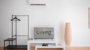 Ein minimalistisches Wohnzimmer mit Fernseher, Kleiderhaken, einem kleinen Tisch und einer hohen Stehlampe.