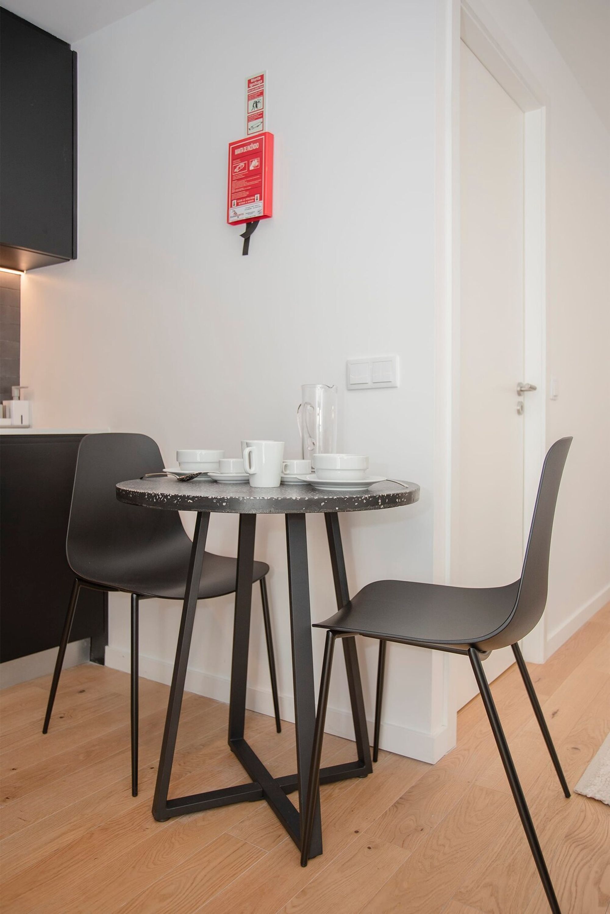 A small dining area with a round table, two black chairs, and a white wall with a red fire safety sign.