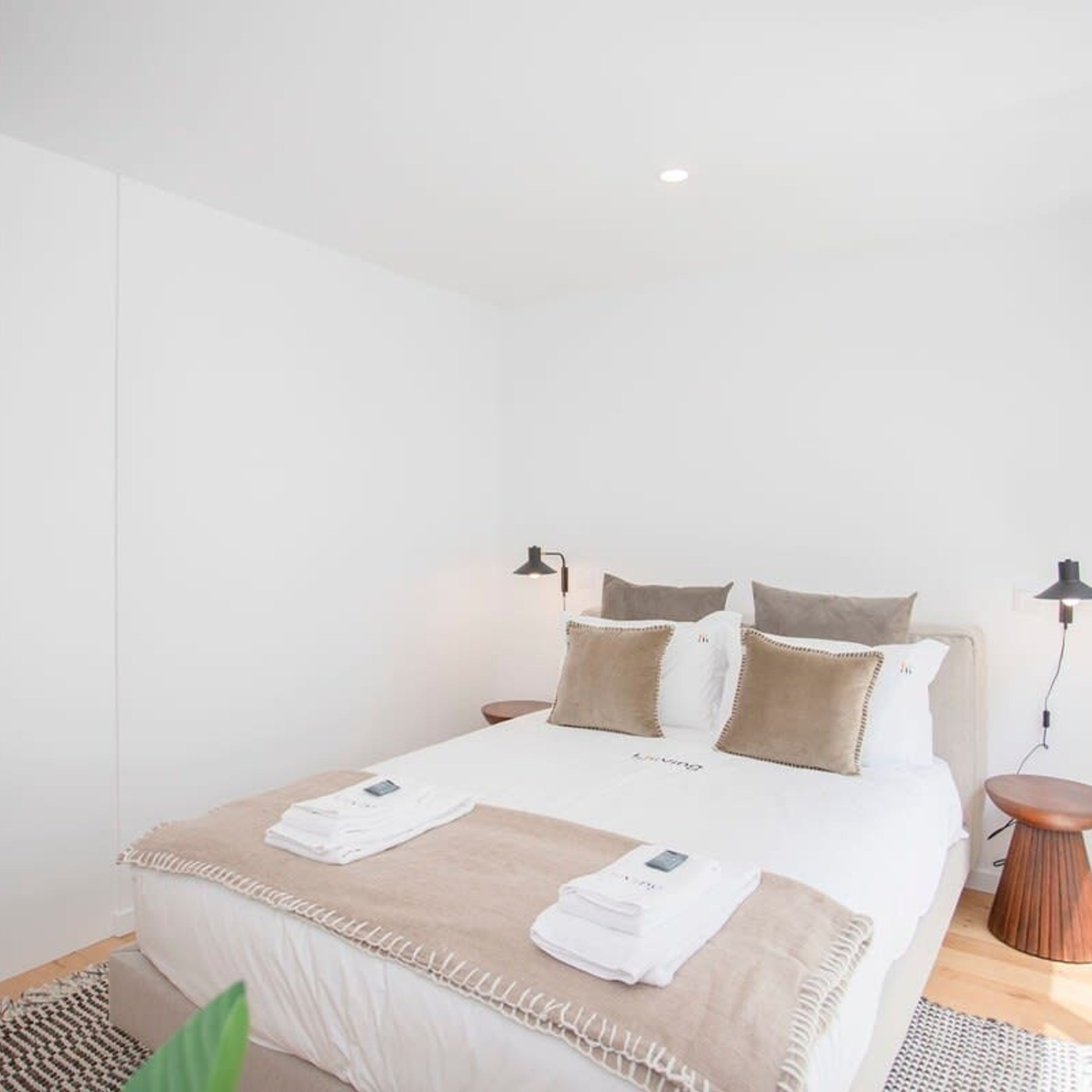 Una habitación de hotel moderna con una cama bien hecha, una mesita de noche, una lámpara y un taburete de madera.