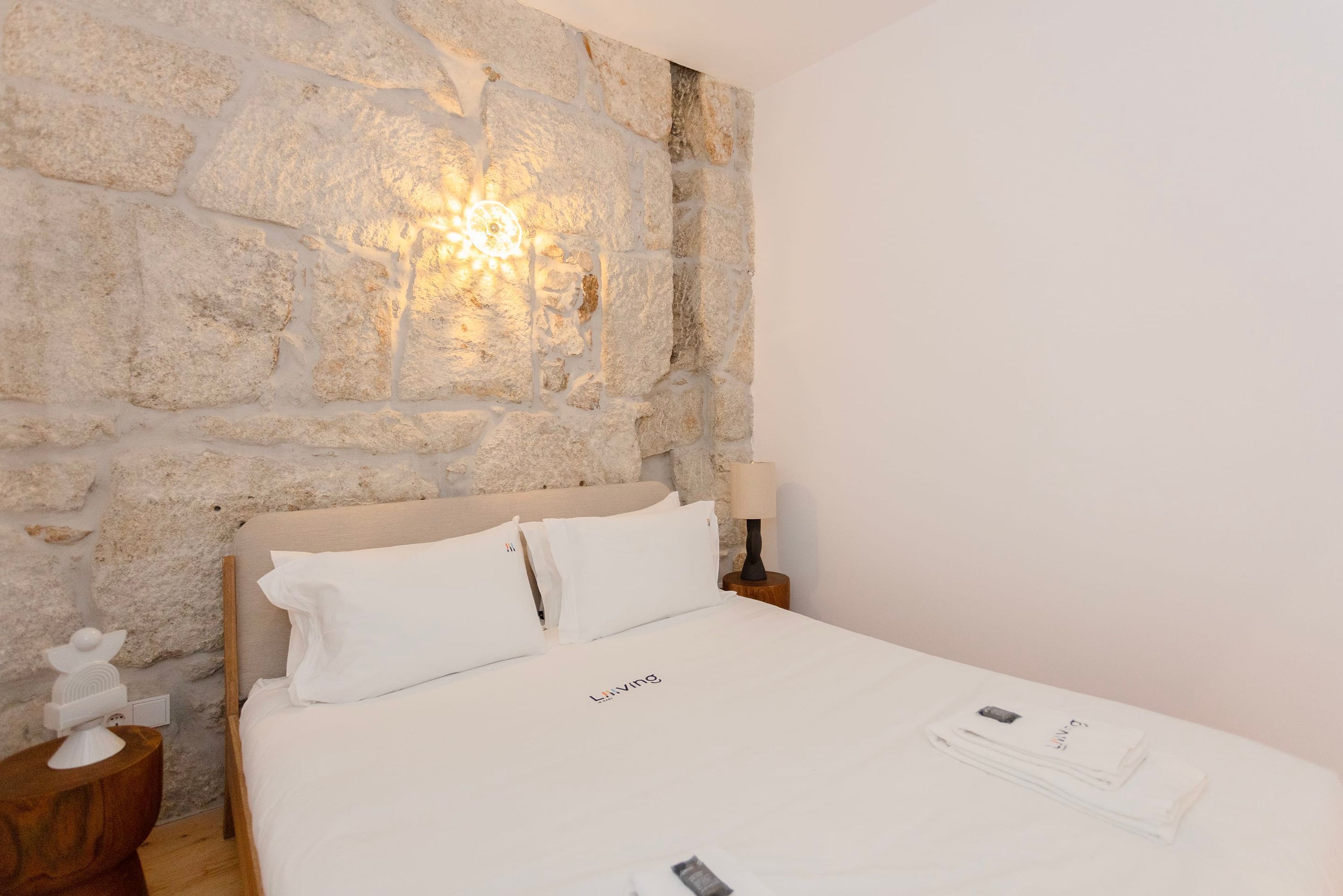 Un lit bien fait, avec du linge de lit blanc et des oreillers, un mur en pierre et une table de chevet avec une lampe.