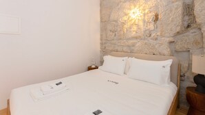 Una cama bien hecha con sábanas y almohadas blancas, una pared de piedra con una lámpara, y una mesita de noche de madera con lámpara.
