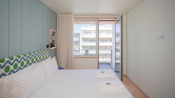 A hotel room with a bed, a window with a view of buildings, a door, and a wall with a patterned headboard. - Allegro Boutique Apartment C by Liiiving (Porto)