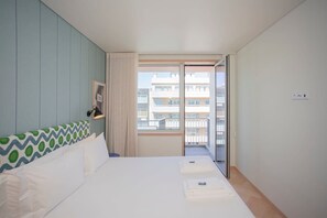 A hotel room with a bed, a window with a view of buildings, a door, and a wall with a patterned headboard. - Allegro Boutique Apartment C by Liiiving (Porto)
