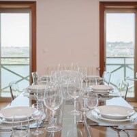 Mesa de comedor preparada con varios platos y copas de vino, ubicada en una habitación con ventanas amplias que dan a un cuerpo de agua.