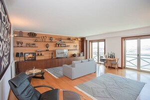 Una sala moderna con un sofá gris, un sillón negro, una pared de madera con varios objetos decorativos, un televisor y una zona de comedor con mesa y sillas.