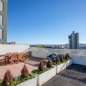 Terraço no telhado com móveis de madeira e vasos, oferecendo vista para a paisagem urbana.