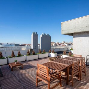 Eine Dachterrasse mit Holztischen und -bänken, umgeben von Pflanzgefäßen mit Sträuchern, mit Blick auf eine Stadtlandschaft mit Gebäuden und blauem Himmel.