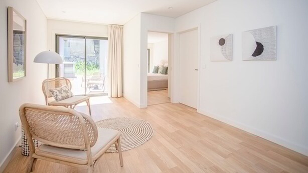 Ein modernes Zimmer mit Holzboden, einem Korbstuhl, einem runden Teppich und Blick durch ein großes Fenster.