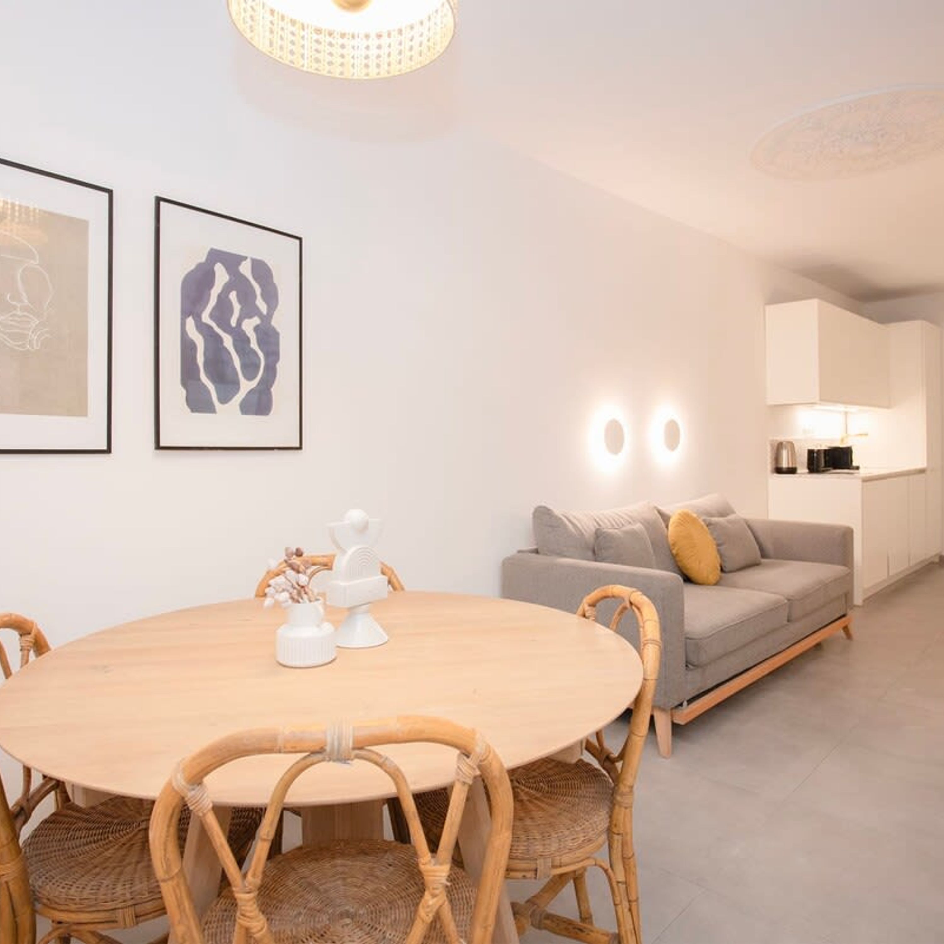 Ein modernes Wohnzimmer mit einem runden Holztisch, Korbstühlen, einem grauen Sofa und einer Küche mit weißen Schränken und einer Lampe.