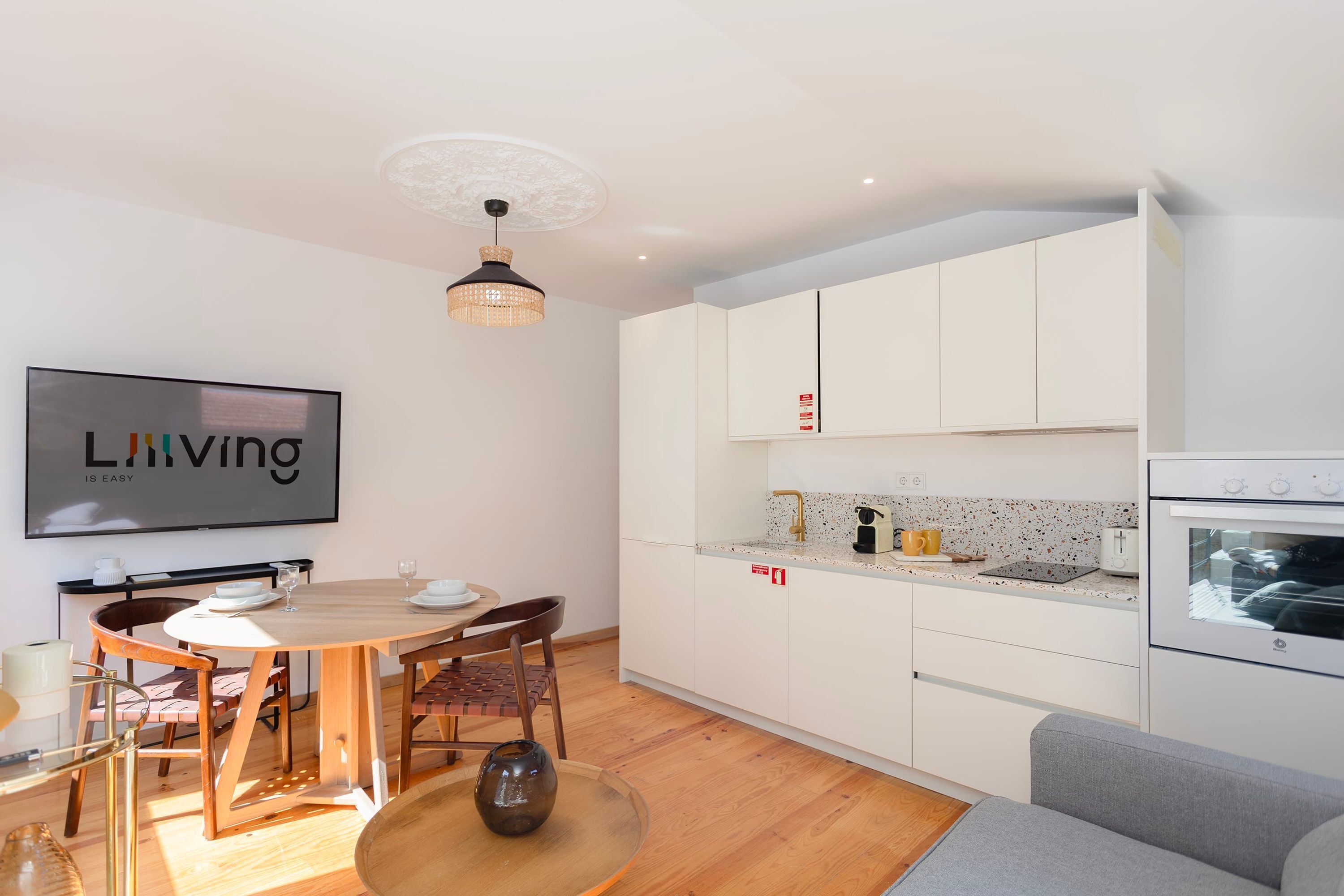 A modern kitchen with white cabinets, a dining table with chairs, and a large TV displaying the text "Living is easy".