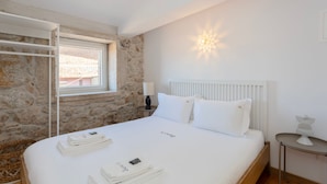 A neatly made bed with white linens and pillows, a stone wall, a window with a view, and a bedside table with a lamp. - Liiiving In Gaia - The Wine Side XVI (Vila Nova de Gaia)