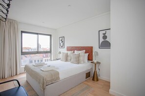 A modern hotel room with a large bed, a view through a window, a bedside table, and a chair. - Liiiving in Porto - City Design Apartment Q (Porto)