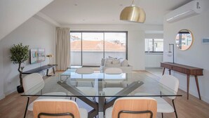 A modern living room with a glass dining table, comfortable seating, a large window with a view, and a ceiling light fixture. - Liiiving in Porto - City Design Apartment Q (Porto)