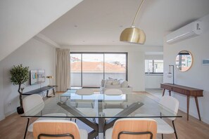 A modern living room with a glass dining table, comfortable seating, a large window with a view, and a ceiling light fixture. - Liiiving in Porto - City Design Apartment Q (Porto)
